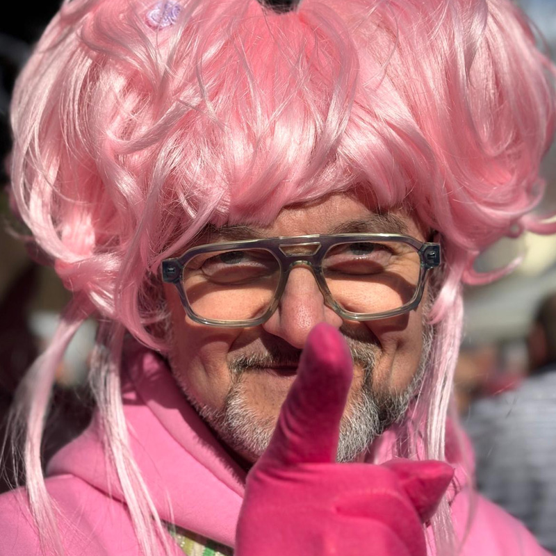 Mann mit Brille und grauem Bart hat eine Rosa Perücke auf einen Rosa Jogginganzug an und zeigt mit auf die Kamera mit seinem pinken Handschuh