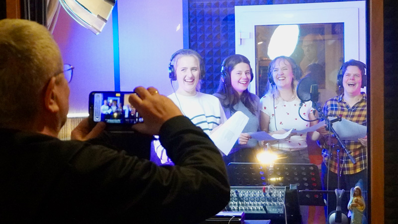 Vier junge Frauen singen heiter im Tonstudio und werden durch die Scheibe von einem Mann mit seinem Smartphone gefilmt