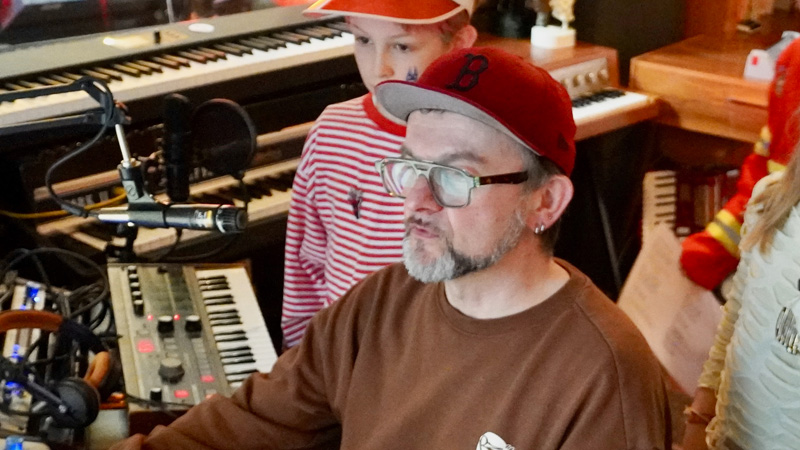 Musikproduzent umringt von kostümierten Kindern schaut auf den Computerbildschirm in seinem Tonstudio