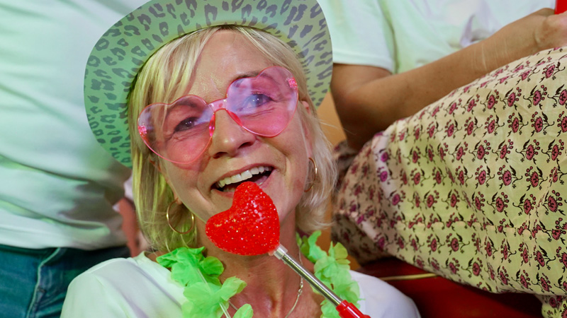 Eine kostümierte Frau mit rosa Herzchensonnenbrille posiert für ein Foto und singt in ein Dekoherz